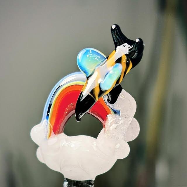 Flameworked glass bee with black and amber stripes riding a rainbow arc on a clear cloud