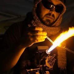 Kyle James Auffrey at a bench torch, holding a glass rod in the flame, wearing dark glasses and a knit cap